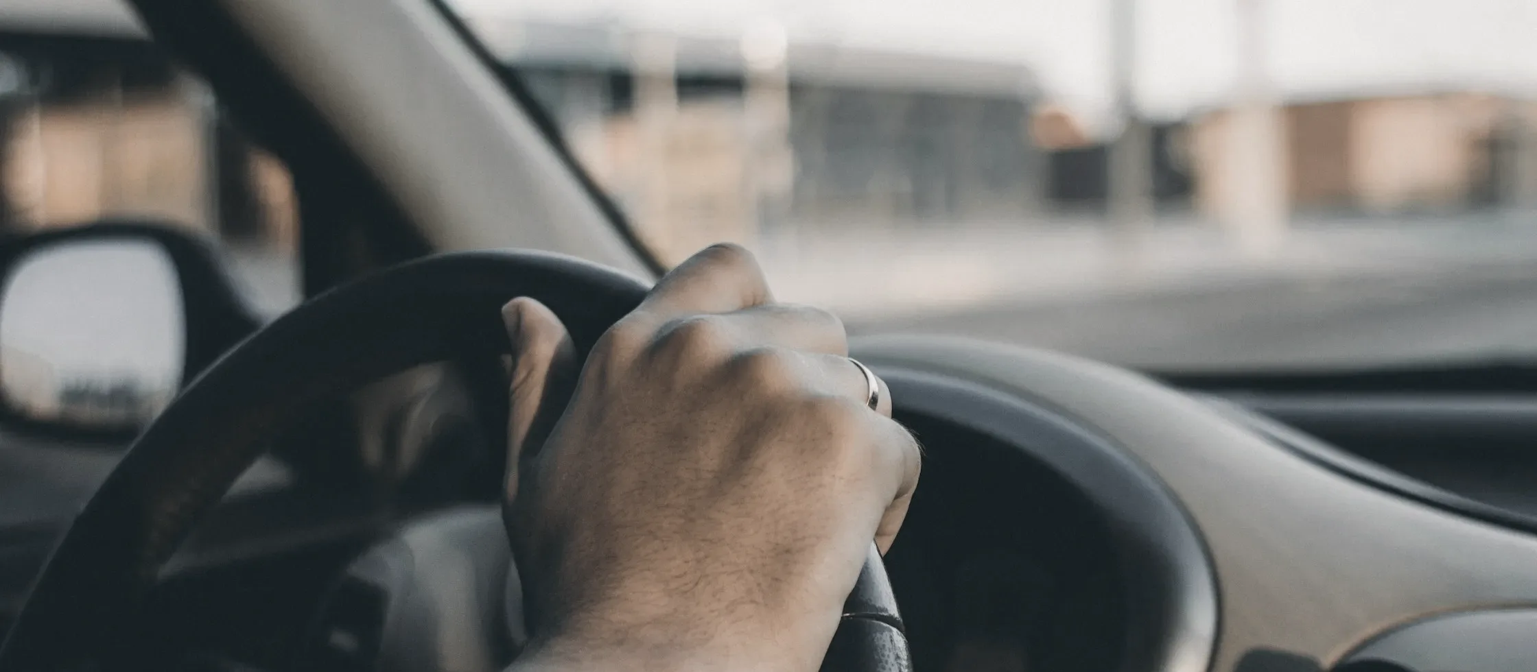 A hand resting on a steering wheel, a low industrial street out of focus through the windscreen.