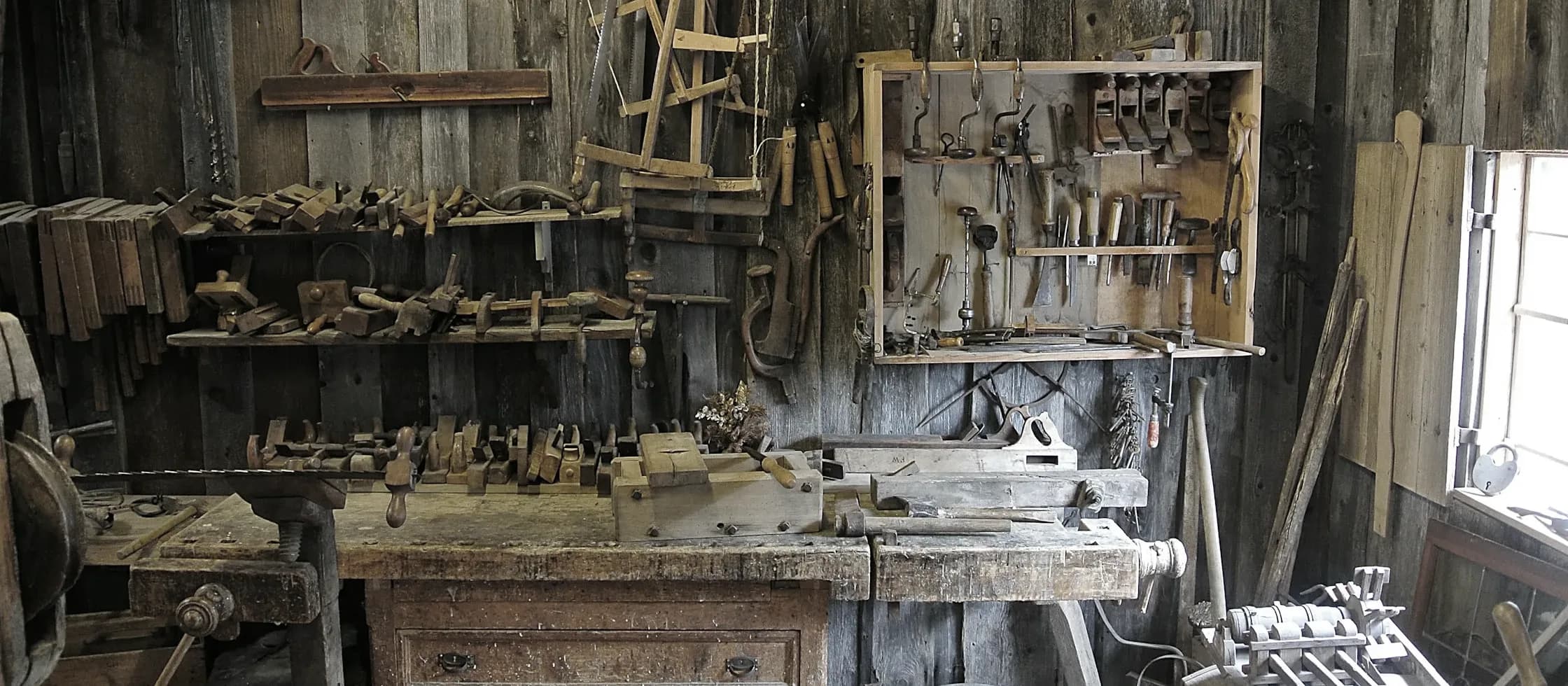 A joiner’s bench against a boarded wall hung with hand planes, saws and braces.